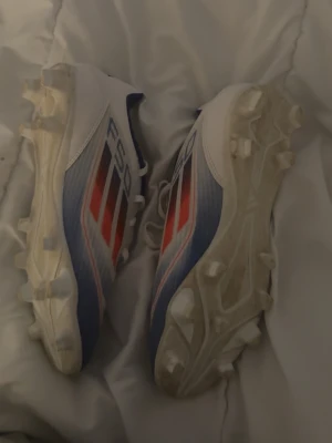 Adidas F50 blåvita fotbollsskor - Säljer ett par Adidas F50 fotbollsskor i blått, vitt och rött. Skorna har klassiska tre ränder på sidan, snörning och dobbar för bra grepp på planen. Tillverkade i syntetmaterial för lätt känsla och snabbhet. Perfekta för dig som vill sticka ut på fotbollsplanen.