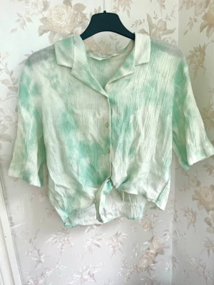 Grönvit skjorta med knyt från H&M - Säljer en luftig grönvit skjorta från H&M med knyt framtill och kort ärm. Skjortan har krage, knappar och ett lätt tyg som känns som linne. Perfekt för en avslappnad och trendig look.