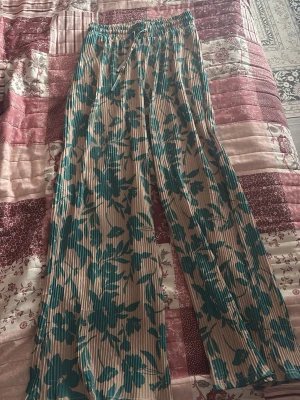 Beige plisserade leggings med blommor - Säljer ett par beige leggings med grönt blommönster och plisserad struktur. De har resår i midjan och raka ben för en avslappnad look. Perfekta för dig som vill ha något bekvämt men ändå trendigt.