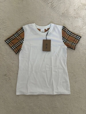 Vit t-shirt med rutiga ärmar Burberry - Snygg vit t-shirt från Burberry med klassiska beige, svarta och röda rutiga kortärmade ärmar. T-shirten har rund halsringning och är tillverkad i mjuk bomull. Perfekt för dig som vill ha en stilren men ändå unik look.