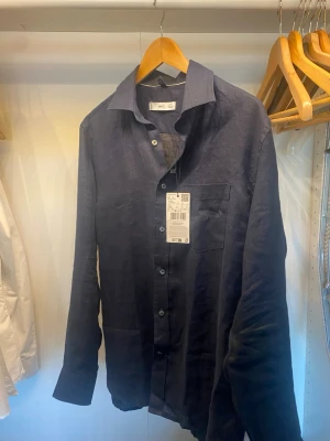 Mörkblå linneskjorta från Arket - Snygg mörkblå skjorta från Arket i 100% linne. Klassisk krage, bröstficka och knäppning framtill. Perfekt för en avslappnad men stilren look. Långärmad modell med normal passform.