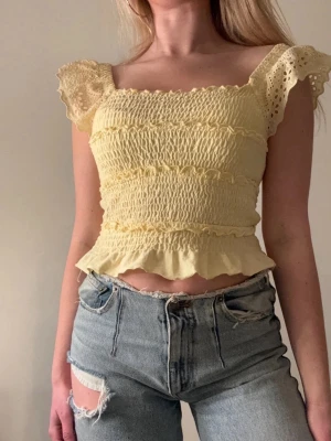 Gul smockad topp med volang - Söt ljusgul smockad topp med volangkant och spetsdetaljer på axelbanden. Croppad modell med fyrkantig ringning och stretchig passform. Perfekt för varma dagar och ger en romantisk vibe till din outfit.