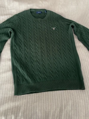 GANT Mörkgrön kabelstickad tröja  - Mörkgrön kabelstickad tröja från Gant med rund halsringning och diskret logga broderad på bröstet. Tröjan har långa ärmar och ribbade muddar vid ärmslut och nederkant. Perfekt för dig som gillar klassisk och stilren look.