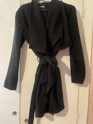 Svart kappa från Fashion Nova - Stilren svart kappa från Fashion Nova med bred krage och knytskärp i midjan. Kappan har lång ärm och en mjuk, något tjockare textil som ger en snygg siluett. Perfekt för dig som vill ha en klassisk och trendig look.