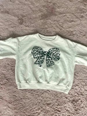 Vit sweatshirt med leopardrosett Lindex - Cool vit sweatshirt från Lindex med en stor rosett framtill i leopardmönster. Tröjan har rund hals, ribbade muddar vid ärmslut och nederkant samt en avslappnad passform. Perfekt för dig som gillar trendiga detaljer och djurmönster.