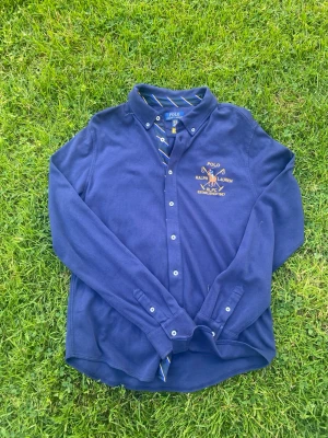 Marinblå skjorta från Polo Ralph Lauren - Perfekt för Sommarkvällarna!Säljer en marinblå långärmad skjorta från Polo Ralph Lauren med broderad gul logga och knappstängning. Skjortan har klassisk krage och detaljer med blå och gula ränder på insidan. Perfekt för dig som gillar stilrena plagg med sportig touch. Size S-M. Skriv till mig för frågor och bud, tveka inte!