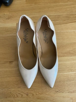 Ljusa klackar med vågig kant - Säljer ett par ljusbeige ballerinaskor från Golden med spetsig tå och unik vågig kant längs öppningen. Skorna är i glansigt material som ger en stilren look. Perfekta för dig som vill ha något extra till outfiten.