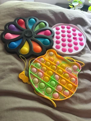 Tre olika pop it-leksaker - Säljer tre pop it-leksaker i olika former och färger. En är rund och vit med rosa bubblor, en är svart med regnbågsfärgade bubblor i blomform och en är gul/grön/orange med rosa och gröna bubblor i form av en katt med snöre.