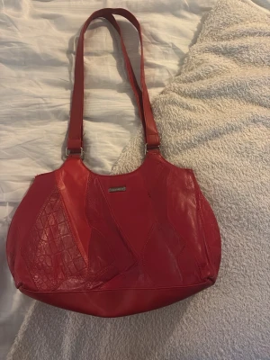 Röd axelremsväska  - Snygg trendig röd handväska i skinn med axelremmar och patchwork-detaljer, där vissa partier har krokopräglat mönster. Väskan har rundad form och metallogo framtill. Perfekt för dig som vill sticka ut med en färgstark accessoar.
