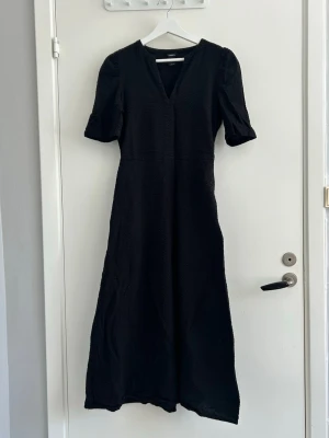 Svart långklänning från Lindex - Svart långklänning från Lindex med v-ringning och korta puffärmar. Klänningen har en strukturerad yta och en enkel, elegant siluett som ger en stilren look. Perfekt för dig som gillar klassiska och tidlösa plagg.