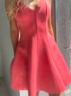 Kort rosa klänning med smala axelband - Snygg kort rosa klänning med v-ringning och smala axelband. Klänningen har en figurnära överdel och utställd kjol som ger en feminin siluett. Perfekt för varma dagar och somriga tillfällen.