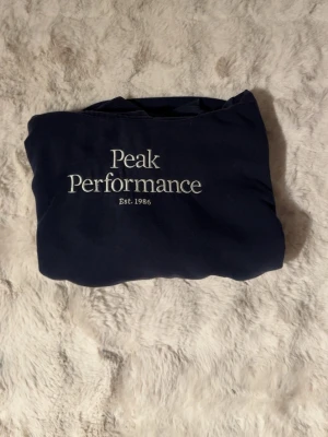 Mörkblå hoodie från Peak Performance - Mörkblå hoodie från Peak Performance med vit broderad logga och texten 'Est. 1986' på bröstet. Klassisk känguruficka framtill och ribbade muddar vid ärmslut och nederkant. Har använts mycket men börjar nu bli för liten, trots användning är den i ett väldigt bra skick. Det är barnstorlek på bilden och i beskrivningen. Pris kan diskuteras (byxorna gå också att köpa)