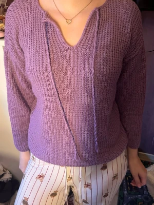 Lila stickad tröja med snörning - Mysig lila stickad tröja med v-ringning och dekorativ snörning framtill. Tröjan har lång ärm och ett ribbat mönster som ger en avslappnad och trendig look. Perfekt att matcha med jeans eller kjol för en chill outfit.