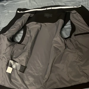 Svart dunväst från Guess - Snygg svart dunväst från Guess med quiltad framsida och hög krage. Västen har två dragkedjefickor framtill och stängs med dragkedja. Materialet är en mix av polyester och syntet, vilket gör den både lätt och bekväm. Perfekt för lager-på-lager-stil. Pris kan diskuteras vid snabb affär.