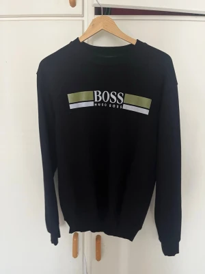 Svart sweatshirt från Hugo Boss - Snygg svart sweatshirt från Hugo Boss med logga och gröna samt vita detaljer på bröstet. Tröjan har rund halsringning och långa ärmar. Perfekt för en stilren och avslappnad look.