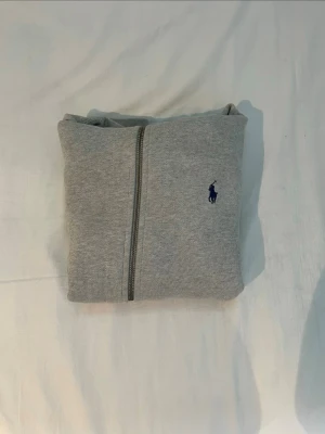 Grå hoodie från Polo Ralph Lauren - Snygg grå hoodie från Polo Ralph Lauren med dragkedja framtill och klassisk blå logga broderad på bröstet. Hoodien har vita dragsnören och mjukt innerfoder. Perfekt för en avslappnad och stilren look.