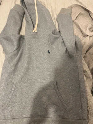 Grå hoodie från Polo Ralph Lauren - Snygg grå hoodie från Polo Ralph Lauren med klassisk blå logga broderad på bröstet. Hoodien har huva med vita snören och en stor magficka framtill. Tillverkad i mjukt sweatshirtmaterial som är perfekt för chill dagar.