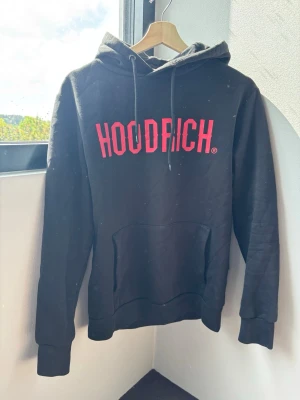 Svart HOODRICH hoodie med röd text - Svart hoodie från HOODRICH med stor röd logga på bröstet. Klassisk modell med huva och dragsnören samt en stor magficka. Tillverkad i mjukt material, perfekt för en chill och avslappnad stil.