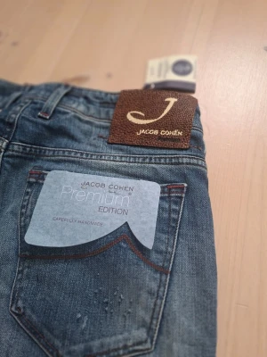 Jacob Cohën Premium Edition jeans - Snygga blå jeans från Jacob Cohën Premium Edition med klassisk femficksmodell och diskreta slitningar. Jeansen har raka ben och bruna detaljer på bakfickan samt ett brunt märke i mockaimitation vid linningen. Perfekta för en trendig och avslappnad look.