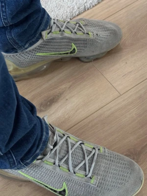Grå Nike Vapormax sneakers med limegrön detalj - Säljer ett par grå Nike sneakers med limegröna detaljer och genomskinlig sula. Skorna har en stickad ovandel och klassisk Nike-logga på sidan. Snygg design med snörning och sportig känsla. Perfekta för dig som gillar streetwear och vill sticka ut.