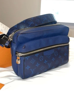 Blå Louis Vuitton axelväska - Äkta Louis Vuitton Messenger väska, köpt i Louis Vuitton-butiken i Paris för 1 460€. Väskan kommer med box, kvitto, certifikat och dustbag.  En stilren modell som sticker ut och är väldigt snygg. Väskan är använd några fåtal gånger men väldigt bra skick. Skick 9/10. Väskan har endast mindre slitage på sidorna av bältet som går runt, men inget som påverkar helhetsintrycket av väskan eller användningen.  Säljer den för endast 7200 kr, bra tillfälle att köpa en exklusiv, fin Louis Vuitton väska.