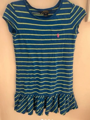 Randig blå kortklänning från Polo - Säljer en blå kortklänning ,barn  från Polo Ralph Lauren med gula ränder och volang nedtill. Klänningen har korta ärmar, rund halsringning och en liten bröstficka med rosa logga. Perfekt för en avslappnad och cool stil. Storlek S , 7år barn