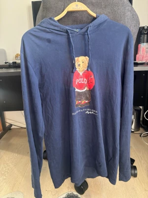 Marinblå hoodie med björntryck - Snygg marinblå hoodie från Polo Ralph Lauren med det klassiska björntrycket på bröstet. Långärmad modell med huva och dragsko. Tillverkad i mjukt och tunt material som känns skönt mot huden. Perfekt för en avslappnad och trendig stil.      Prister ej hugget i sten