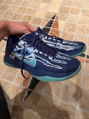 Kobe 5 xray - Unika blå Nike Kobe sneakers med coolt skelettmönster på ovandelen och ljusblå sula. Skorna har lågt skaft, klassisk snörning och är tillverkade i syntetmaterial och textil. Perfekta för dig som vill sticka ut med din sneakerstil.