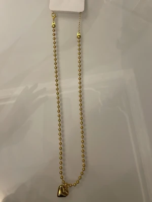 Guldfärgat halsband med hjärthänge - Snyggt halsband i guldfärg med små pärlor längs hela kedjan och ett hjärtformat hänge. Perfekt accessoar för att lyfta din outfit med en trendig touch. Halsbandet har justerbar längd och stängs med karbinhake.