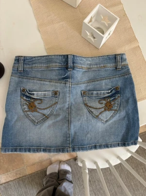 Ljusblå jeanskjol med broderade fickor - Säljer en ljusblå jeanskjol i kort modell med klassiska fem fickor och bälteshällor. Kjolen har snygga broderade detaljer på bakfickorna och stängs med dragkedja och dubbla knappar framtill. Perfekt till sommaren och enkel att matcha.