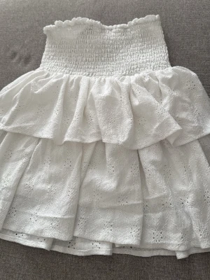 Vit volangkjol med broderade detaljer - Söt vit kortkjol med bred smock i midjan och dubbla volanger. Kjolen har fina broderade detaljer i tyget. Perfekt för varma dagar och enkel att matcha med olika toppar.