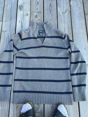 Gråblå randig stickad tröja Polo - Zipen är borta Stickad tröja i grått med mörkblå horisontella ränder från Polo Ralph Lauren. Tröjan har en hög krage med dragkedja och den klassiska röda loggan broderad på bröstet. Långa ärmar och ribbade muddar vid ärmslut och nederkant.OBS barnstorlek 