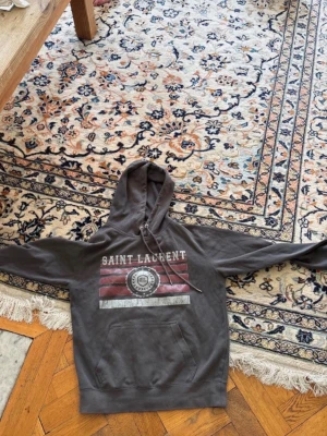 Mörkgrå Saint Laurent hoodie - Säljer en mörkgrå hoodie från Saint Laurent med tryck framtill i vitt och rött. Hoodien har en klassisk huva med dragsko och en stor magficka. Perfekt för dig som gillar streetwear och vill ha något stilrent men ändå med attityd.