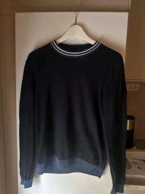 Mörkblå HUGO BOSS tröja med randig krage - En stilren mörkblå tröja med långa ärmar och ribbade muddar. Kragen har en sportig look med vita och ljusblå ränder. Perfekt för dig som gillar en enkel men snygg stil. Materialet känns mjukt och passar bra till jeans eller chinos.