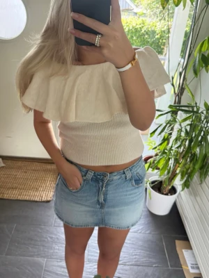 Beige croppad topp med volang - Trendig croppad topp i beige med stor volang runt axlarna. Toppen är ribbad och har en offshoulder-design som ger en snygg och modern look. Perfekt att matcha med jeans eller kjol för en cool outfit.