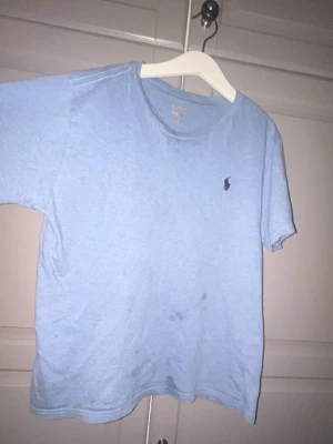 Ljusblå t-shirt från Polo Ralph Lauren - Ljusblå t-shirt från Polo Ralph Lauren med klassisk rund hals och liten broderad logga på bröstet. Skön passform och tillverkad i mjuk bomull, perfekt för en avslappnad stil.