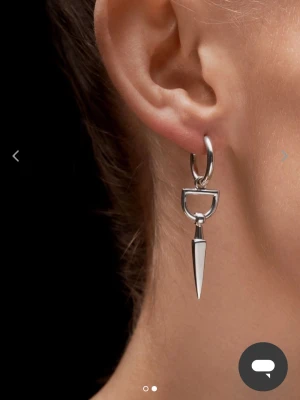Maria Nilsdotter örhänge - Coolt örhänge i silverfärgad metall med en unik design. Örhänget har en rund ögla och en hängande, spetsig detalj som ger en edgy och modern känsla. Perfekt för dig som vill sticka ut med dina accessoarer.