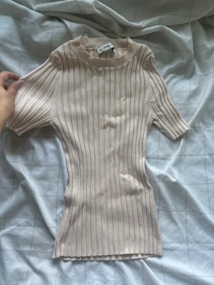 Beige ribbad topp från Zara - Säljer en beige ribbad topp från Zara med korta ärmar och hög hals. Toppen har en slim passform och är tillverkad i ett mjukt, stretchigt material som sitter snyggt på kroppen. Perfekt att styla med jeans eller kjol för en clean look.