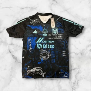 Adidas Tigres UANL svart/blå matchtröja - Säljer en Adidas Tigres UANL matchtröja i svart med blå och vita grafiska detaljer. Tröjan har tryckta loggor och sponsorer på både fram- och baksidan, samt ljusblå Adidas-ränder på axlarna. Materialet är lätt polyester och designen är modern med sportig känsla.