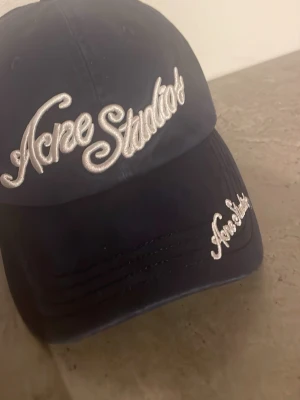 Mörkblå keps från Acne Studios - Snygg mörkblå keps från Acne Studios med vit broderad logga framtill och på skärmen. Klassisk böjd skärm och justerbar passform bak. Tillverkad i bomull för en bekväm känsla och stilren look.