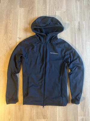 Mörkblå Zip-up hoodie från Peak Performance - Snygg mörkblå hoodie från Peak Performance med dragkedja hela vägen, två fickor med dragkedja och diskret logga på bröstet. Tillverkad i mjukt och stretchigt material som känns skönt mot huden. Perfekt för dig som gillar en clean och sportig stil.