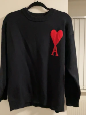  Ami Paris tröja  - Svart stickad tröja från Ami Paris med röd hjärtlogga och bokstaven A på bröstet. Tröjan har rund halsringning, långa ärmar och ribbade muddar vid ärmslut och nederkant. Perfekt för dig som gillar stilrena plagg med en twist.