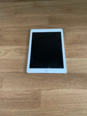 Apple iPad 6th  - Snygg iPad från Apple med vit ram runt skärmen och en stilren silverfärgad baksida med Apple-loggan. Surfplattan har en stor touchskärm och rundade hörn, perfekt för streaming, surf och appar.