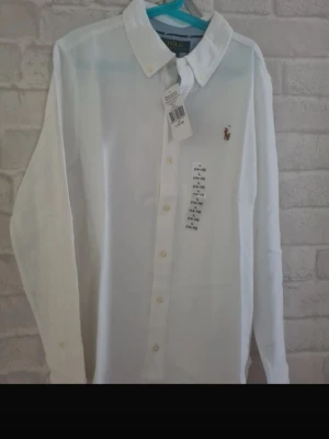 Vit skjorta från Polo Ralph Lauren - Klassisk vit skjorta från Polo Ralph Lauren med broderad logga på bröstet. Skjortan har lång ärm, knappar framtill och en stilren krage. Perfekt för en clean och tidlös look.