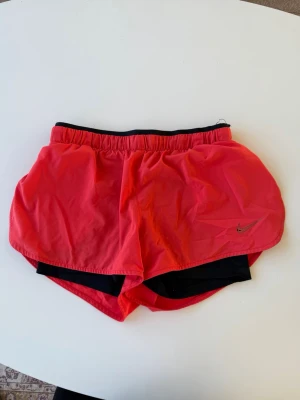 Röda/ orange löparshorts från Nike - Snygga korall färgade löparshorts från Nike med svart innerbyxa och elastisk midja. Perfekt för träning och löpning. 