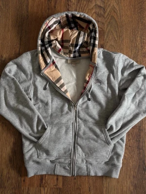 Grå hoodie från Burberry med dragkedja - Storlek L men passar mer som M! Säljer en grå hoodie från Burberry med klassiskt rutigt foder i huvan. Tröjan har dragkedja framtill, två fickor och ribbade muddar. Perfekt för dig som gillar stilrena plagg med en lyxig touch. Ikoniskt Burberry-märke broderat på bröstet.