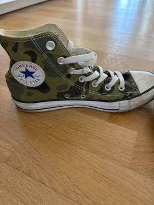 Converse All Star camo high tops - Säljer ett par Converse All Star sneakers i klassisk high top-modell med grön camo-mönstrad canvas och vita snören. Skorna har vit gummisula med svart rand och Converse-logga på sidan. Perfekta för dig som vill sticka ut med en streetig stil. Något olika nyans på gummit 