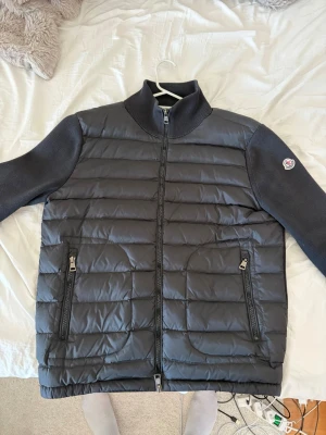 Svart pufferjacka från Moncler - Snygg svart pufferjacka från Moncler med quiltad front och rygg, samt stickade ärmar och krage. Jackan har två dragkedjeförsedda fickor framtill och Moncler-logga på vänster ärm. Perfekt för kalla dagar och riktigt stilren look.