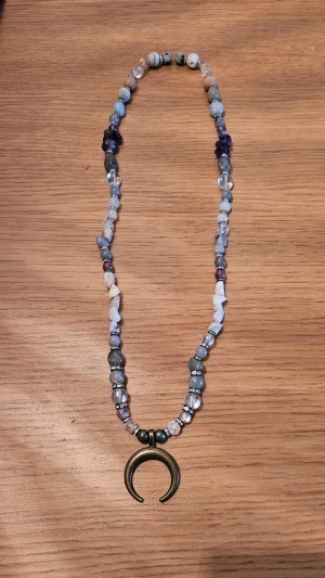 Krafthalsband med halvädelstenar och halvmåne - Unikt halsband med blandade pärlor av halvädelstenar i blått, vitt och lila samt en stor halvmåneformad berlock i metall. Pärlorna är av olika former och material, vilket ger ett coolt och bohemiskt uttryck. Perfekt accessoar för att lyfta din outfit. Detta är det perfekta halsbandet för dig som är intresserade av kristaller och deras magiska egenskaper. Jag gör dessa smycken själv och halsbandet kommer i en söt liten ask tillsammans med information om kristallernas magiska egenskaper.
