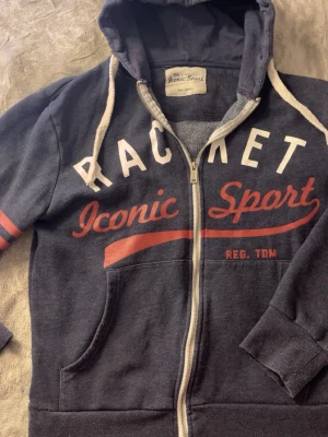 Mörkblå hoodie Lager 157 - Mörkblå hoodie med dragkedja från Lager 157. Tryck i vitt och rött med texten 'Racket Iconic Sport' på bröstet. Två fickor fram, vita snören i huvan och röda ränder på ärmen. Mjuk bomullsblandning och ribbade muddar. Lite nopprig, se bild. 
